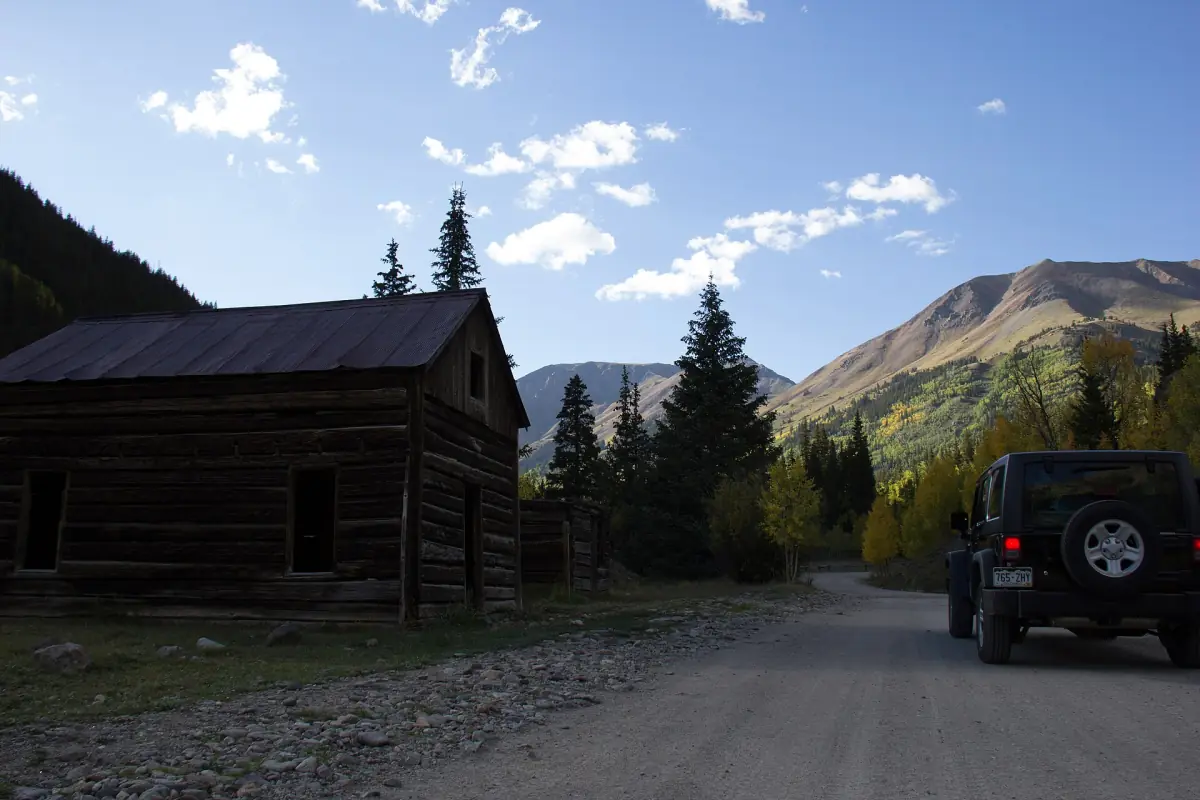 A weathered log cabin beside the dirt road through Animas Forks in autumn, with a parked Jeep and golden aspens on the slopes (BLM photo, September 2013).