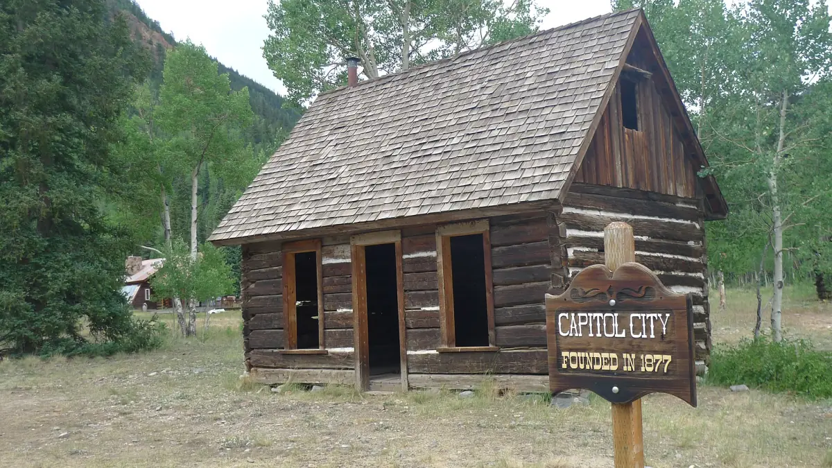 Summer view of a log cabin with a shingle roof next to the 'Capitol City, Founded in 1877' sign on the Alpine Loop.